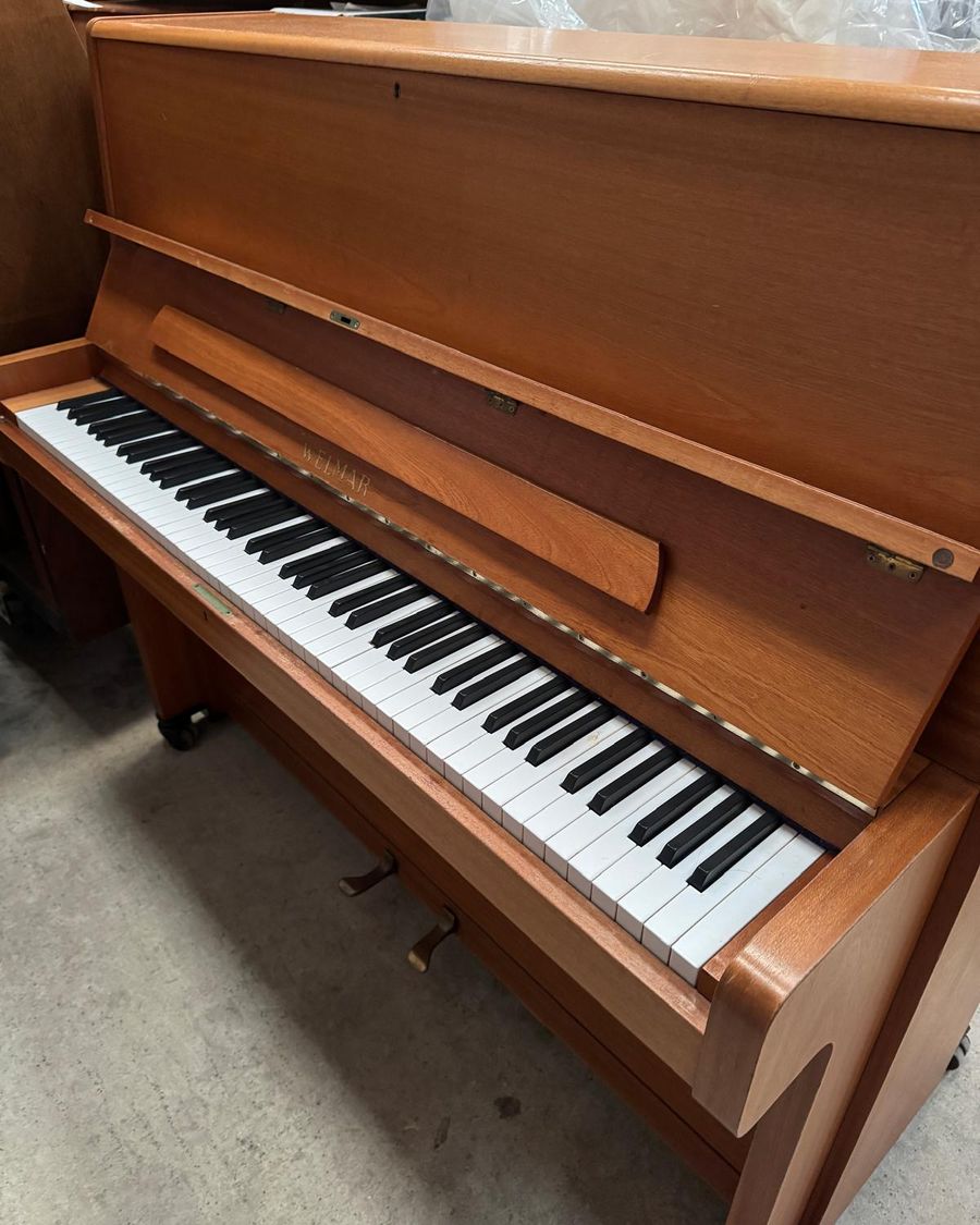 A teak-cased Welmar upright piano on castors in the Piano Rental storage unit, keyboard lid open